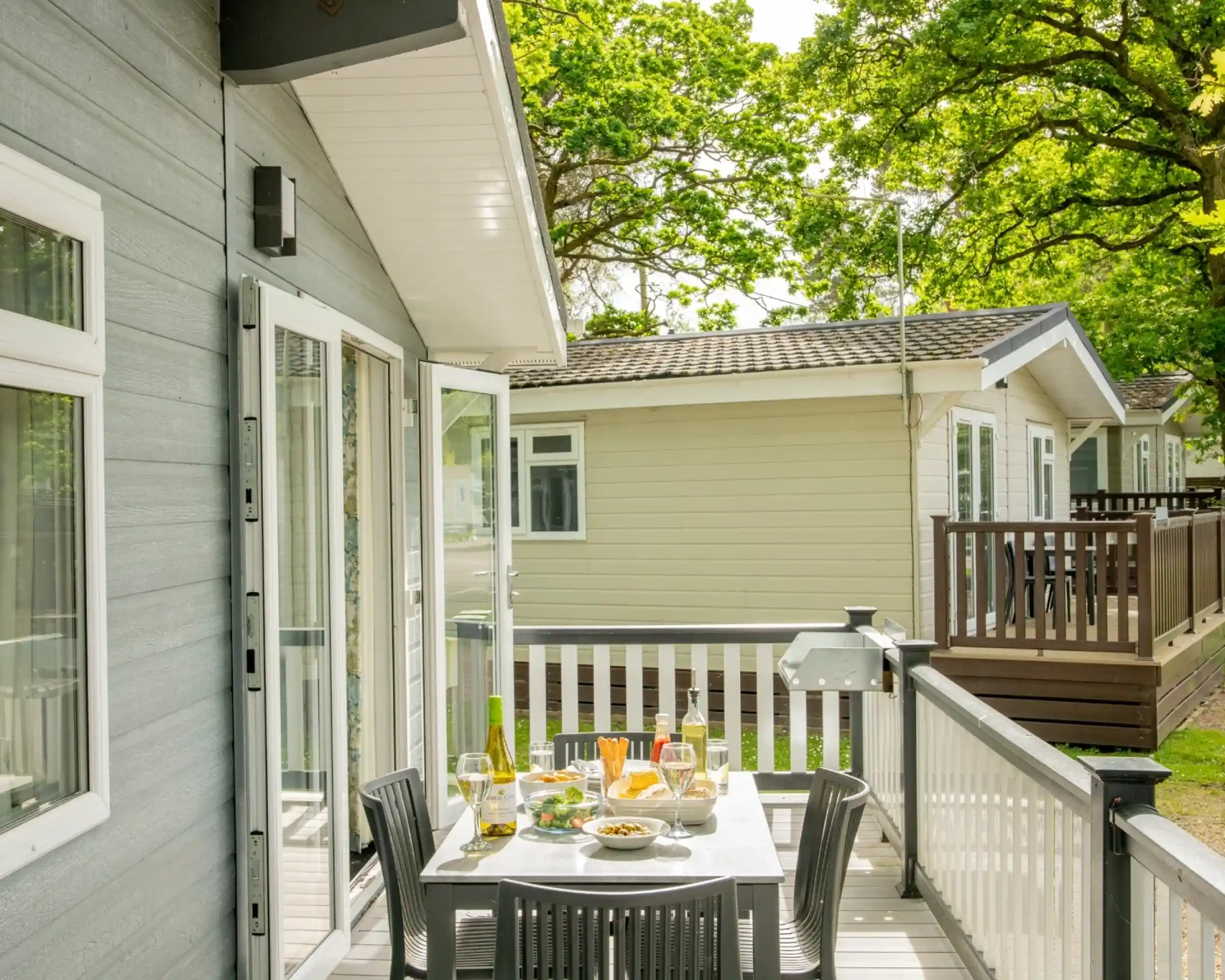 A wooden deck with a table set for dining, featuring snacks and drinks. In the background, two cosy vacation cabins are visible, surrounded by lush green trees and sunlight.