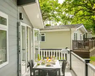 A wooden deck with a table set for dining, featuring snacks and drinks. In the background, two cosy vacation cabins are visible, surrounded by lush green trees and sunlight.
