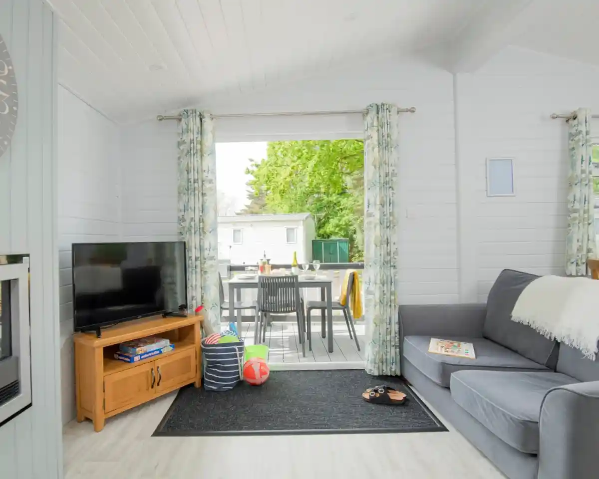 A bright and airy living space featuring a grey sofa, a wooden coffee table with books, and a flat-screen TV. Large windows with floral curtains provide a view of an outdoor dining area. The room has light-coloured walls and a cosy rug.