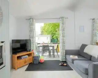 A bright and airy living space featuring a grey sofa, a wooden coffee table with books, and a flat-screen TV. Large windows with floral curtains provide a view of an outdoor dining area. The room has light-coloured walls and a cosy rug.