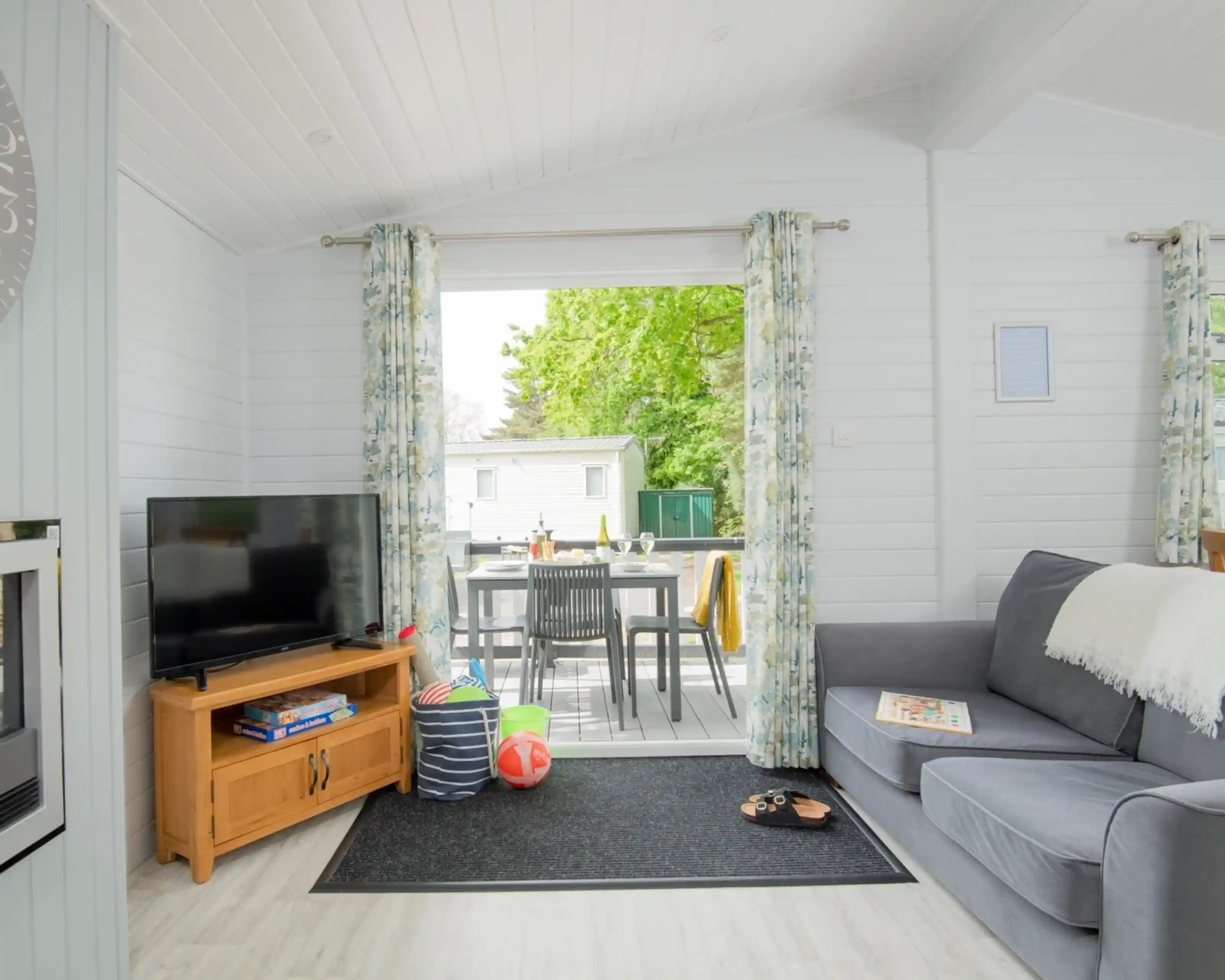 A bright and airy living space featuring a grey sofa, a wooden coffee table with books, and a flat-screen TV. Large windows with floral curtains provide a view of an outdoor dining area. The room has light-coloured walls and a cosy rug.