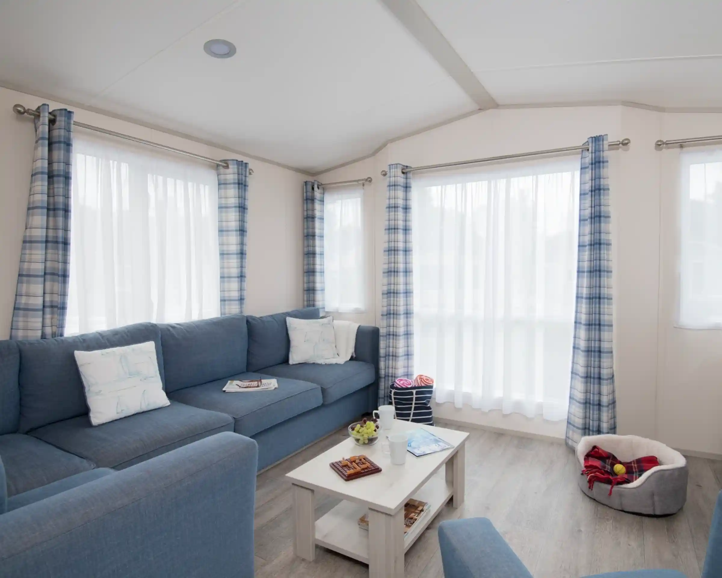 A cosy living room with a blue couch adorned with decorative pillows. A small white coffee table holds magazines and snacks. Light filters through sheer white curtains between blue and white checked drapes. A plush pet bed is visible in the corner.