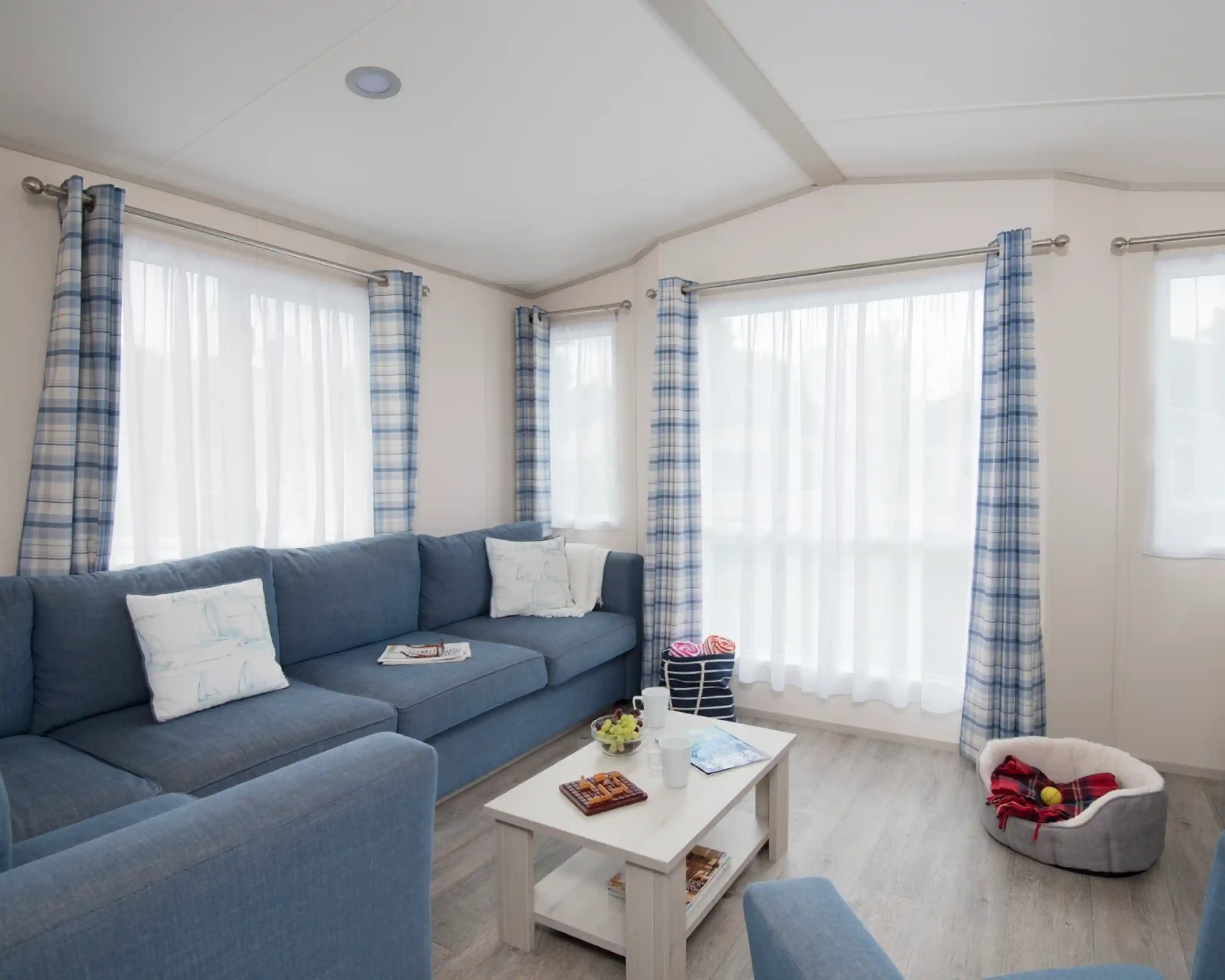 A cosy living room with a blue couch adorned with decorative pillows. A small white coffee table holds magazines and snacks. Light filters through sheer white curtains between blue and white checked drapes. A plush pet bed is visible in the corner.