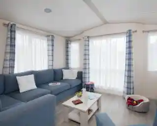 A cosy living room with a blue couch adorned with decorative pillows. A small white coffee table holds magazines and snacks. Light filters through sheer white curtains between blue and white checked drapes. A plush pet bed is visible in the corner.