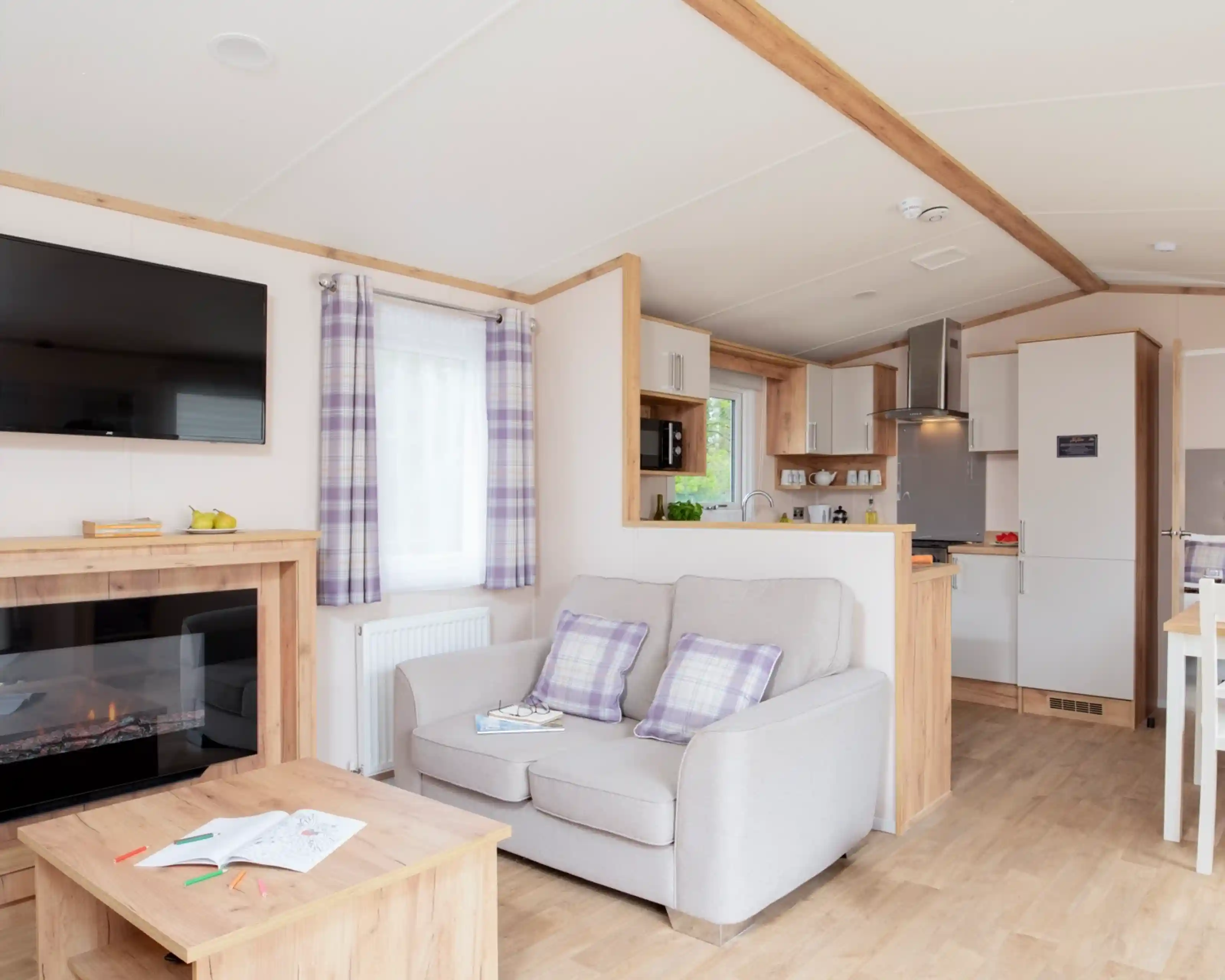 A cosy living area featuring a light grey sofa with plaid cushions, a wooden coffee table with open books, a mounted TV, and a small kitchenette with modern appliances. Warm wood accents and natural light create a welcoming atmosphere.