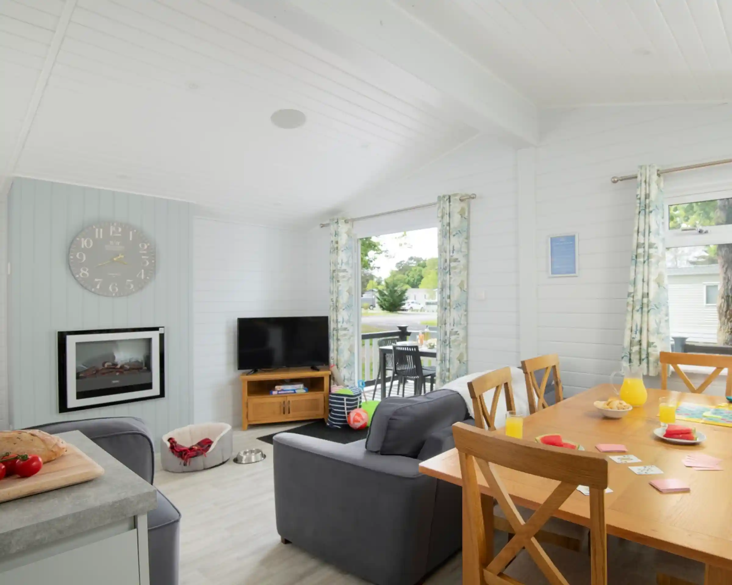 A bright, modern living space featuring a cosy dining area with a wooden table set for breakfast. A plush grey sofa is in the foreground, and a TV is mounted on the wall. Large windows let in natural light, and the room has light-coloured wooden panelling. Decorations include a clock, curtains with floral patterns, and a small outdoor seating area visible through the windows.