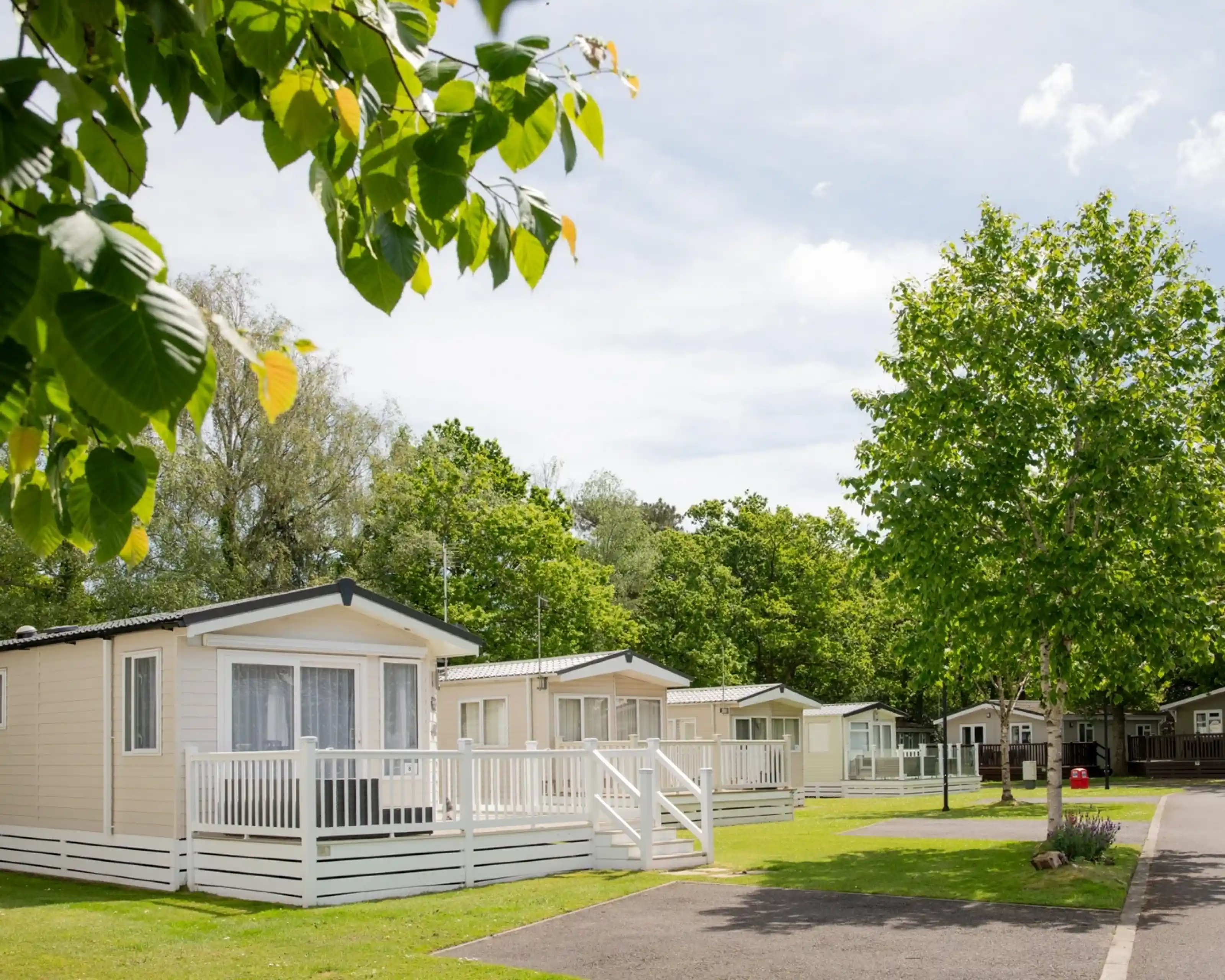 A row of vacation cabins with porches, surrounded by lush green trees and grass. The sky is clear with scattered clouds, creating a peaceful outdoor setting.