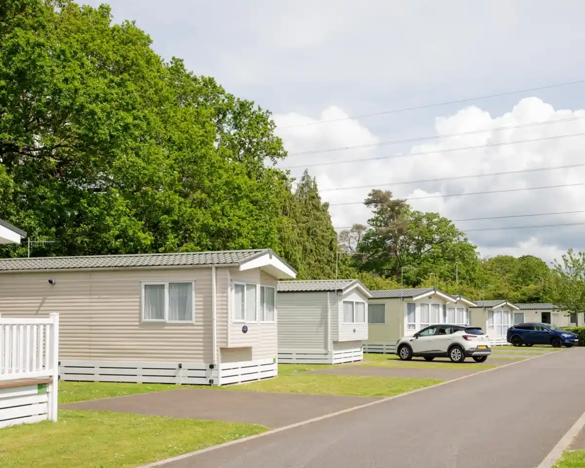 Rows of neatly arranged holiday homes with greenery in the background. A paved road runs alongside the homes, which have white trim and light-coloured siding. Fluffy clouds are visible in the blue sky.