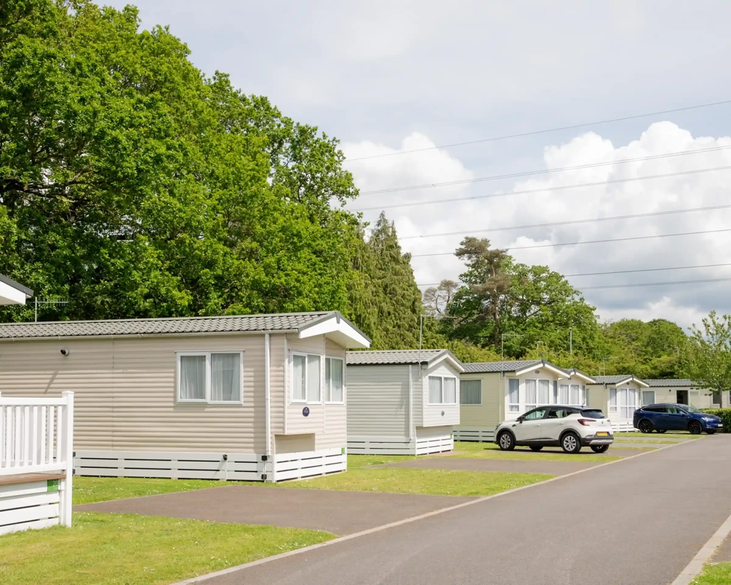Rows of neatly arranged holiday homes with greenery in the background. A paved road runs alongside the homes, which have white trim and light-coloured siding. Fluffy clouds are visible in the blue sky.