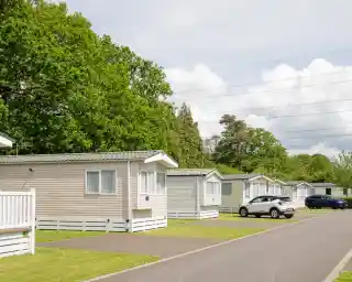 Rows of neatly arranged holiday homes with greenery in the background. A paved road runs alongside the homes, which have white trim and light-coloured siding. Fluffy clouds are visible in the blue sky.