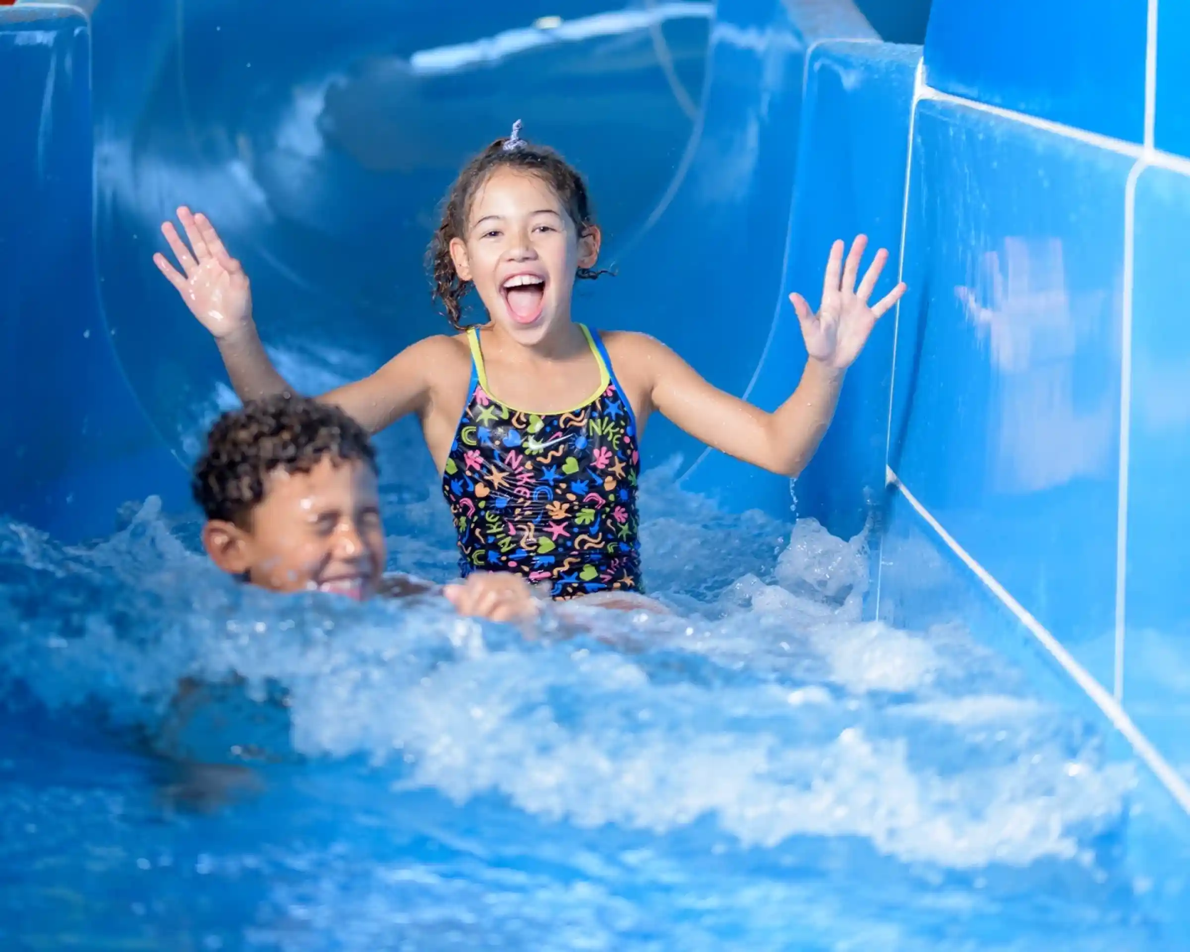 A girl in a colourful swimsuit joyfully slides down a water slide, splashing water around her. A boy swims happily behind her, both enjoying a day at the water park.