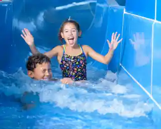 A girl in a colourful swimsuit joyfully slides down a water slide, splashing water around her. A boy swims happily behind her, both enjoying a day at the water park.