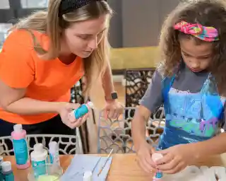 A woman in a bright orange shirt assists a young girl with curly hair, wearing a colourful headband, as they engage in a creative project involving paint and supplies at a table.