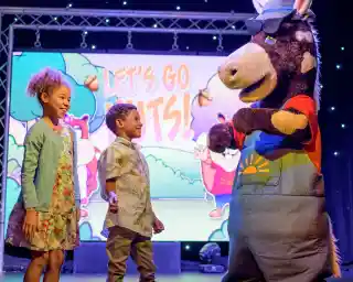 A cheerful mascot dressed as a donkey entertains two smiling children on stage. The backdrop features colourful animated graphics with the text Let's Go Nuts!