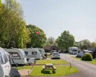 A scenic camping site featuring several caravans and motorhomes lined along a tree-lined pathway. A picnic table is visible in the foreground, and a red flag flies on a pole in the background under a clear blue sky.