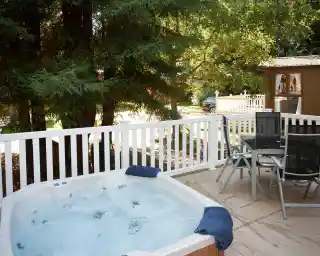 A small hot tub with bubbles is located on a wooden deck, surrounded by tall trees. A table and chairs are set nearby, with a white railing framing the space. A cabin can be seen in the background.