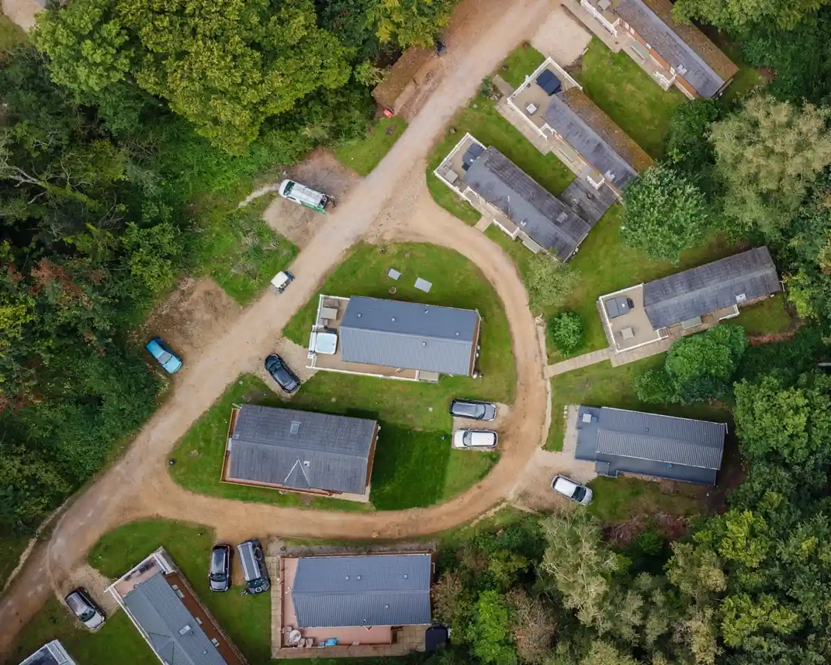 Aerial view of a residential area featuring several single-story buildings arranged around a curved road. Green grass and trees surround the homes, with parked cars visible along the pathway.