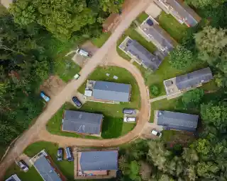 Aerial view of a residential area featuring several single-story buildings arranged around a curved road. Green grass and trees surround the homes, with parked cars visible along the pathway.