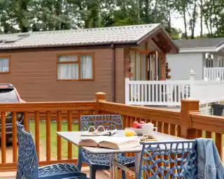 A wooden deck features a table set with an open book, a small plate of snacks, a cup, and a glass of juice. Two blue chairs are nearby, with a serene caravan park in the background surrounded by trees.