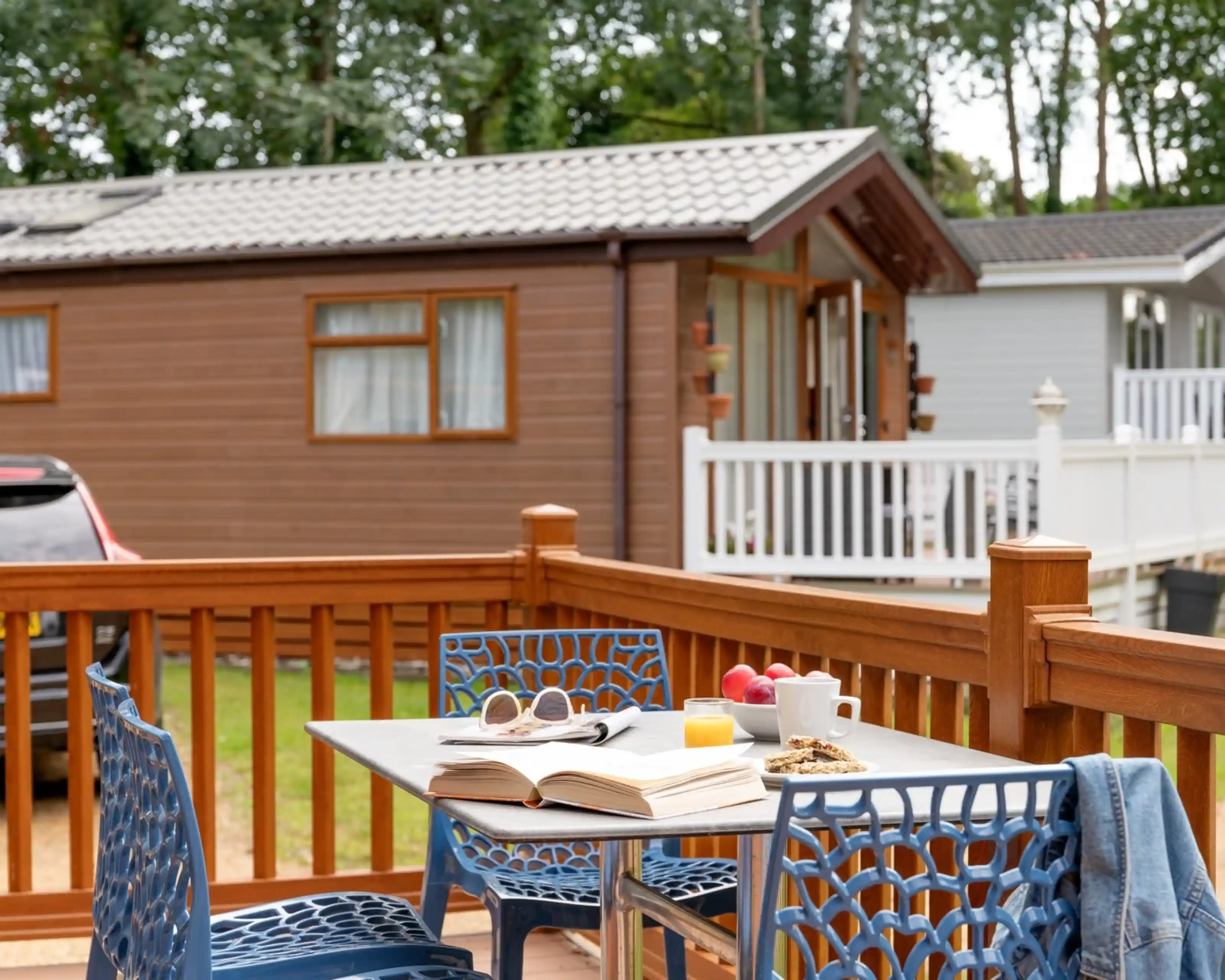 A wooden deck features a table set with an open book, a small plate of snacks, a cup, and a glass of juice. Two blue chairs are nearby, with a serene caravan park in the background surrounded by trees.
