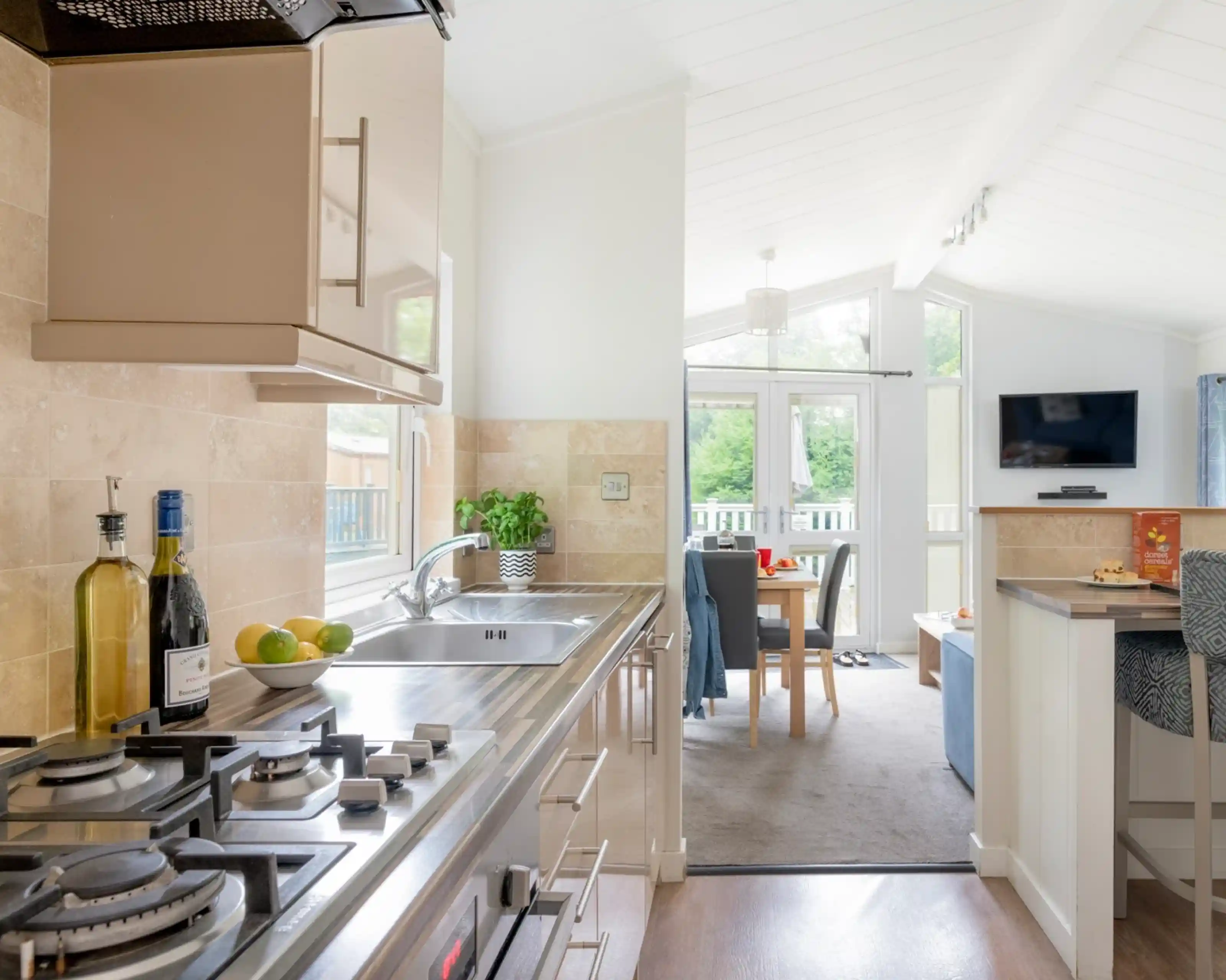 Modern kitchen with sleek appliances, including a gas stove and a double sink. The cabinetry features a light wood finish, complemented by a small dining area visible in the background. Bright natural light floods the space through large windows, creating a welcoming atmosphere.