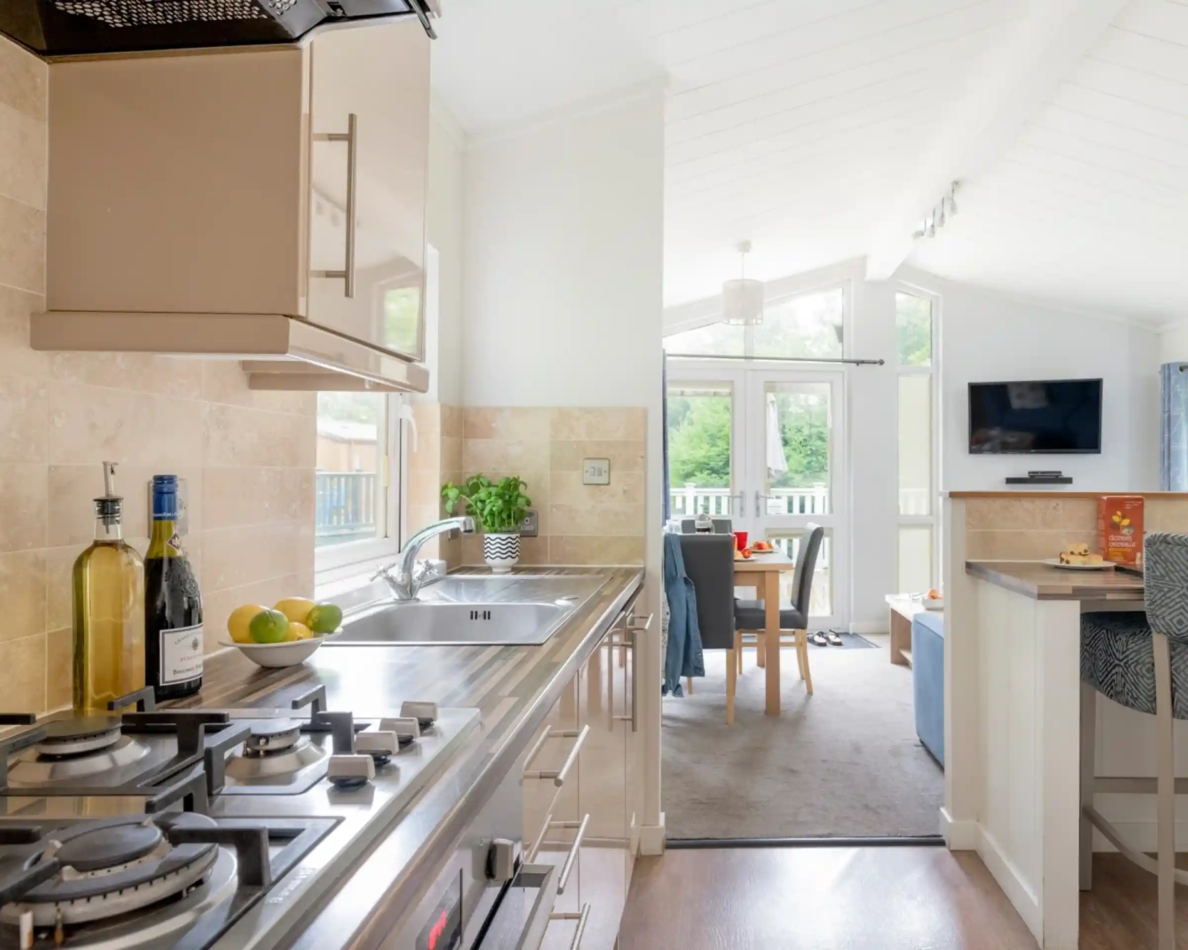 Modern kitchen with sleek appliances, including a gas stove and a double sink. The cabinetry features a light wood finish, complemented by a small dining area visible in the background. Bright natural light floods the space through large windows, creating a welcoming atmosphere.