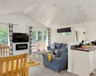 Bright and airy modern living room with a blue sofa, a wooden dining table, and a flat-screen TV mounted on the wall. Large windows let in natural light, and a small kitchen area is visible in the background. Cosy décor includes a striped curtain and a plate of snacks on the table.