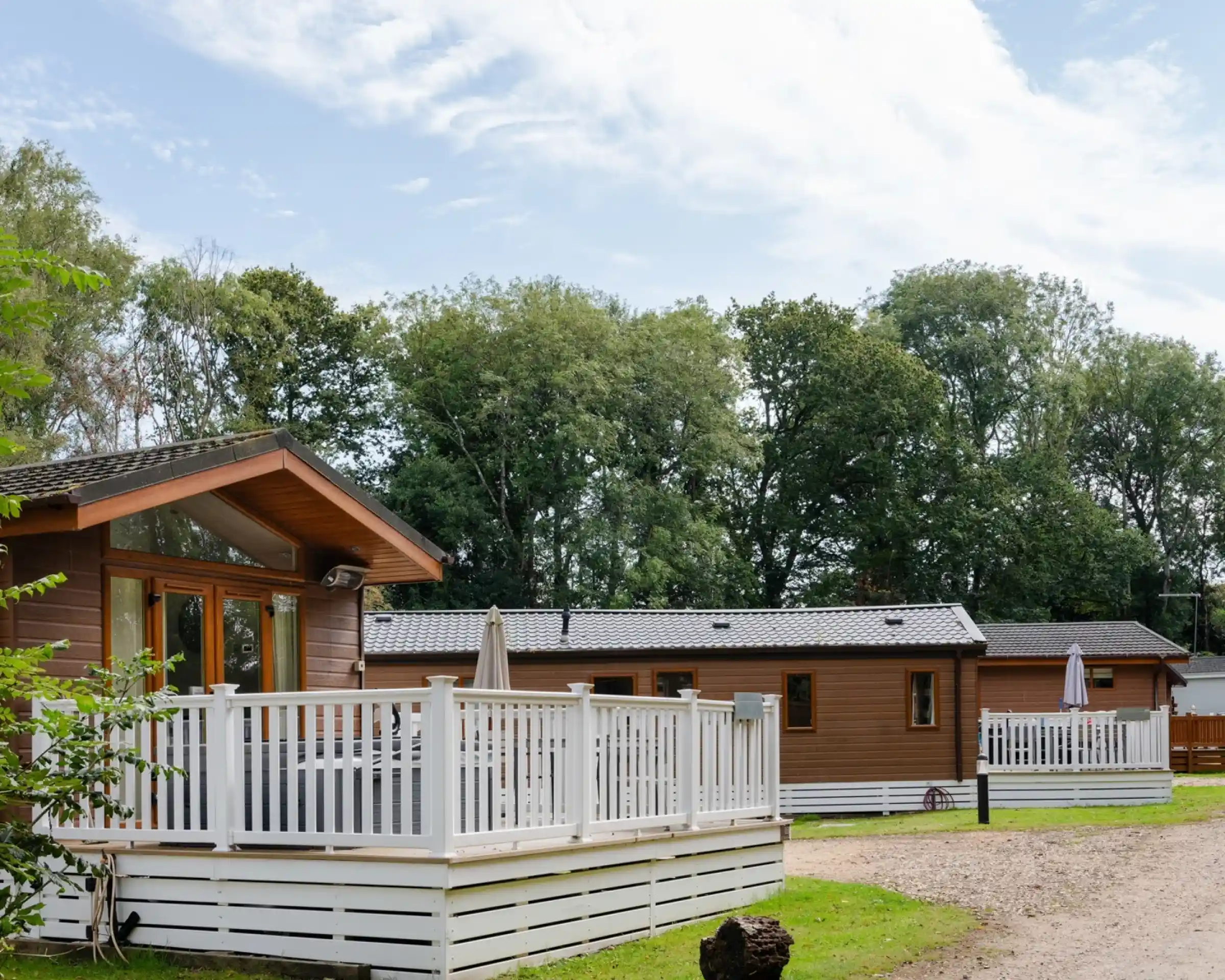 Charming wooden lodges with white railings situated along a gravel path, surrounded by lush green trees and a blue sky.