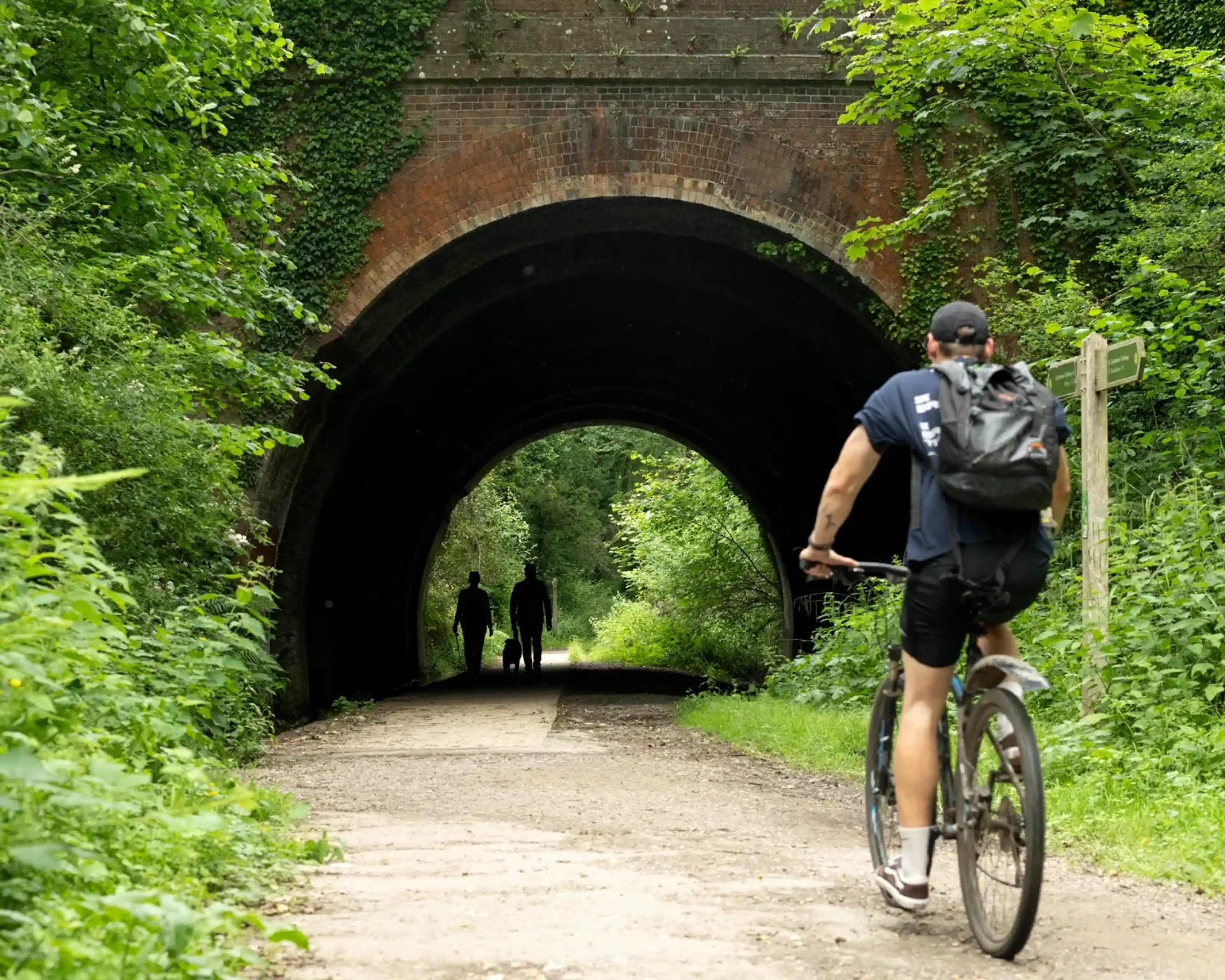 A cyclist rides along a pathway leading into a dark tunnel, where two silhouettes of pedestrians can be seen walking towards the light at the other end. Lush greenery flanks the trail.