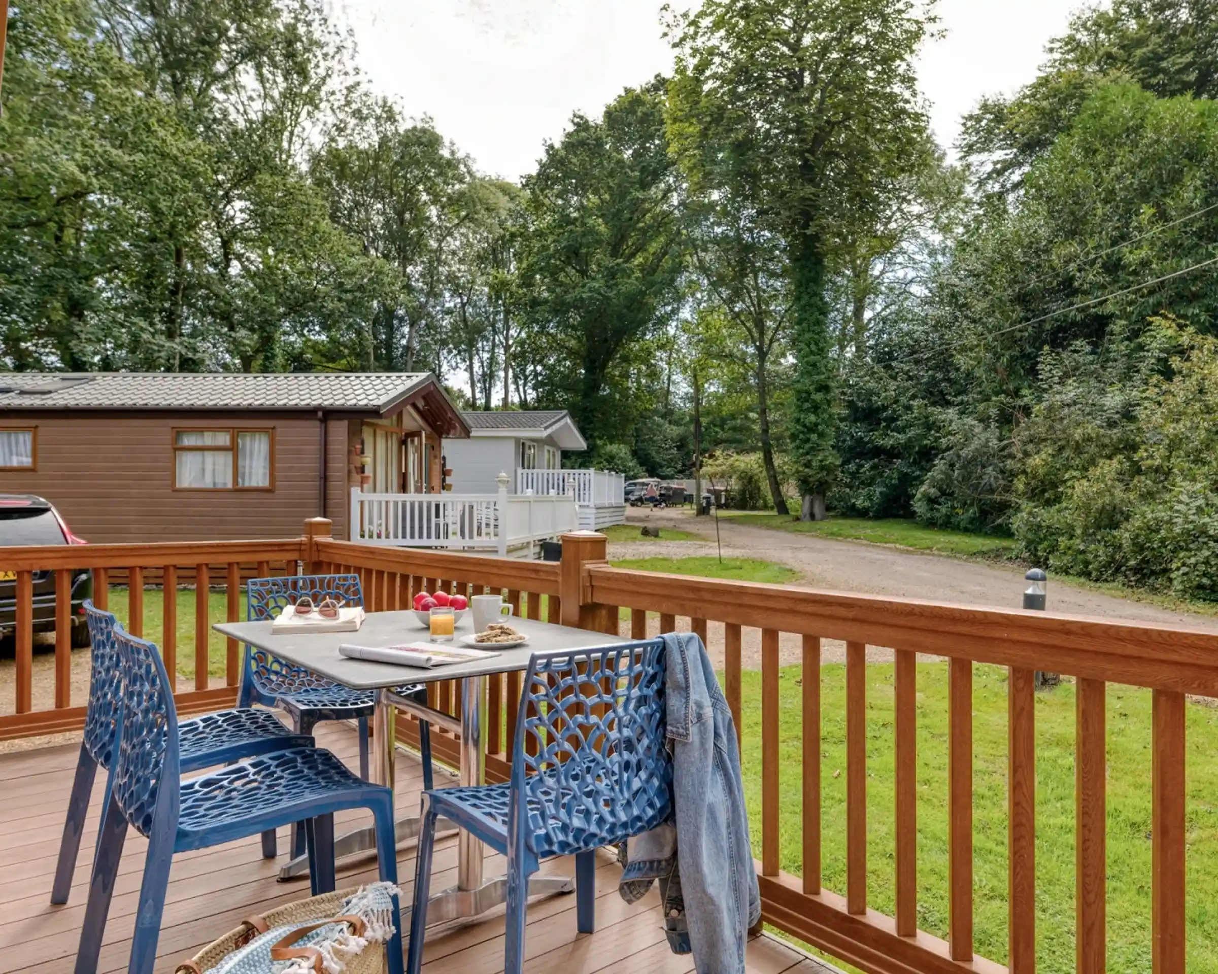 A cosy outdoor deck features a table set for two with plates of breakfast and a denim jacket draped over a chair. In the background, there are cabins surrounded by lush trees and a gravel path.