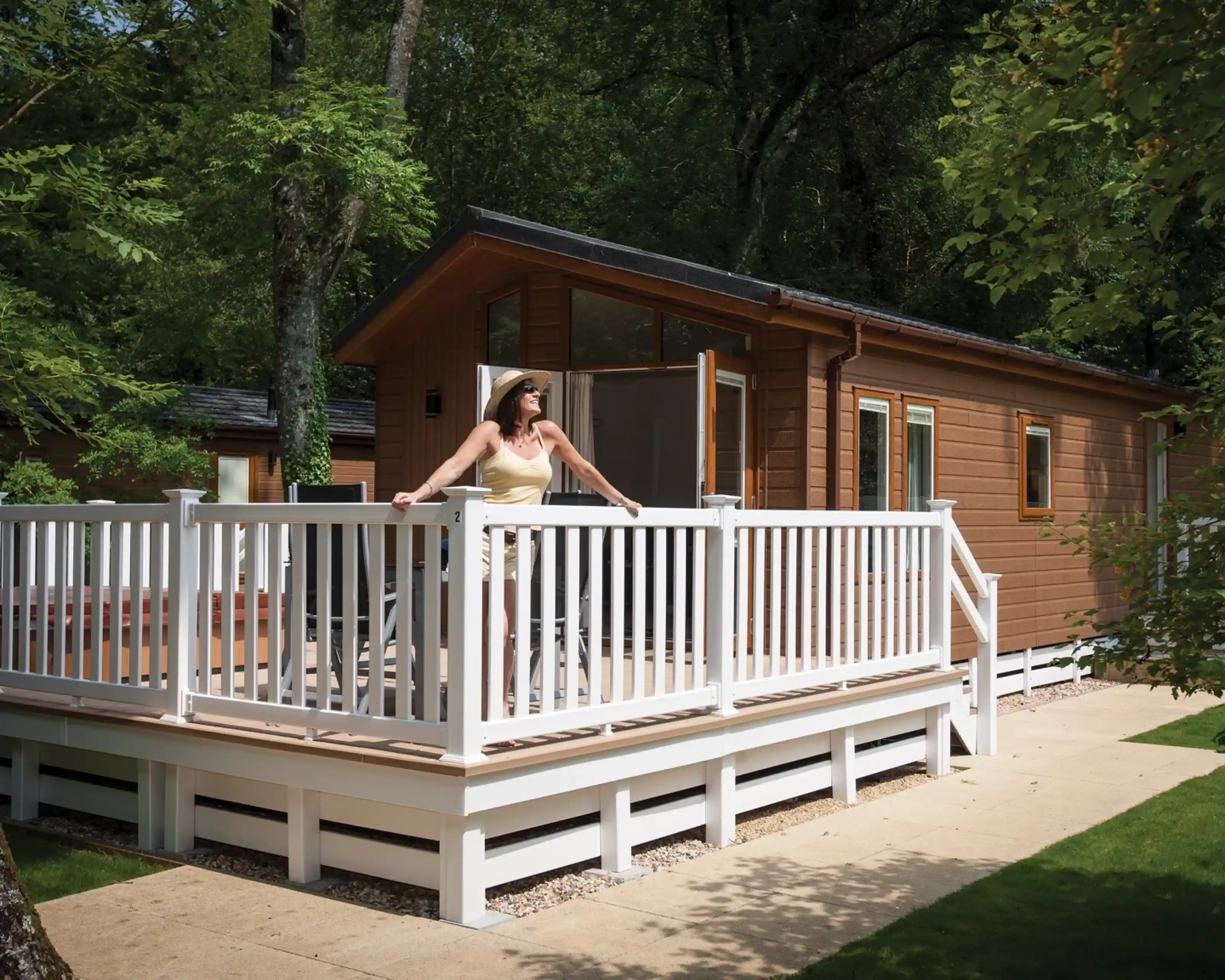 A woman stands on a wooden deck in front of a cabin, surrounded by lush greenery. The cabin features large windows and a warm brown exterior. She wears a light outfit and a hat, enjoying the outdoor setting.
