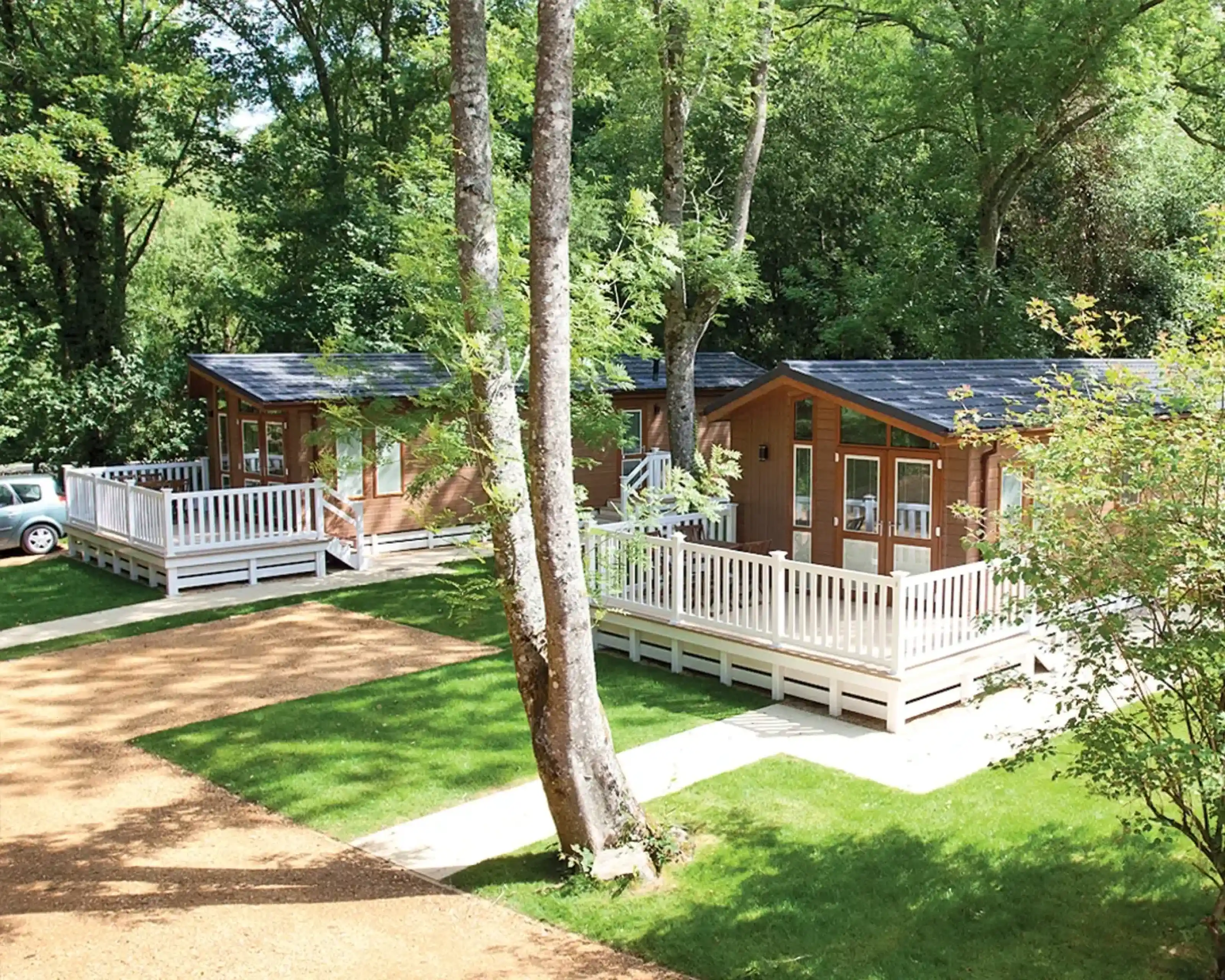 Two wooden cabins with porches are nestled among lush green trees. The cabins have large windows and are situated on a grassy area, with a gravel path leading up to them. A car is parked nearby.