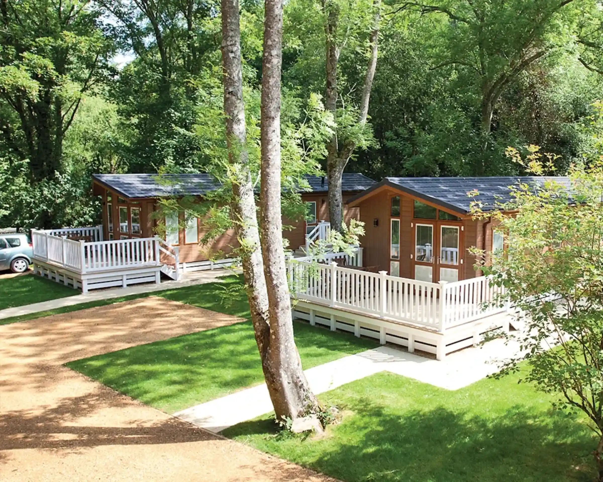 Two wooden cabins with porches are nestled among lush green trees. The cabins have large windows and are situated on a grassy area, with a gravel path leading up to them. A car is parked nearby.