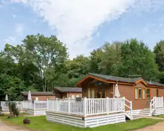 Cozy wooden cabins with white decks surrounded by lush greenery and trees under a partly cloudy blue sky.
