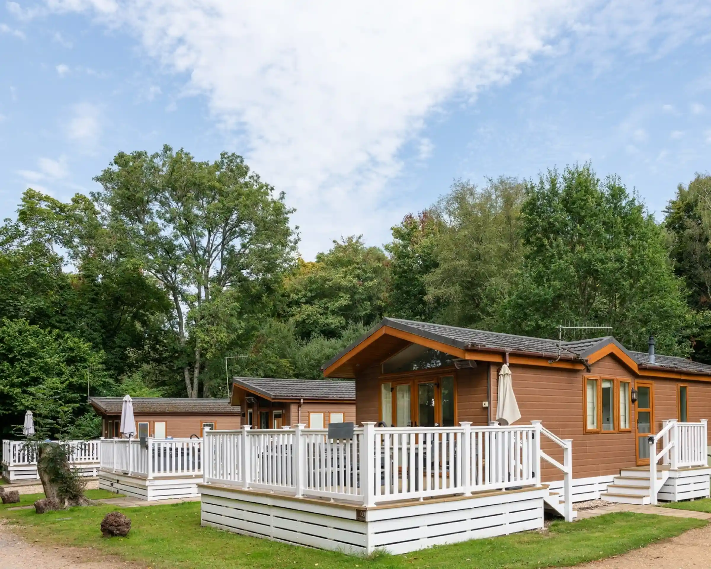 Cozy wooden cabins with white decks surrounded by lush greenery and trees under a partly cloudy blue sky.