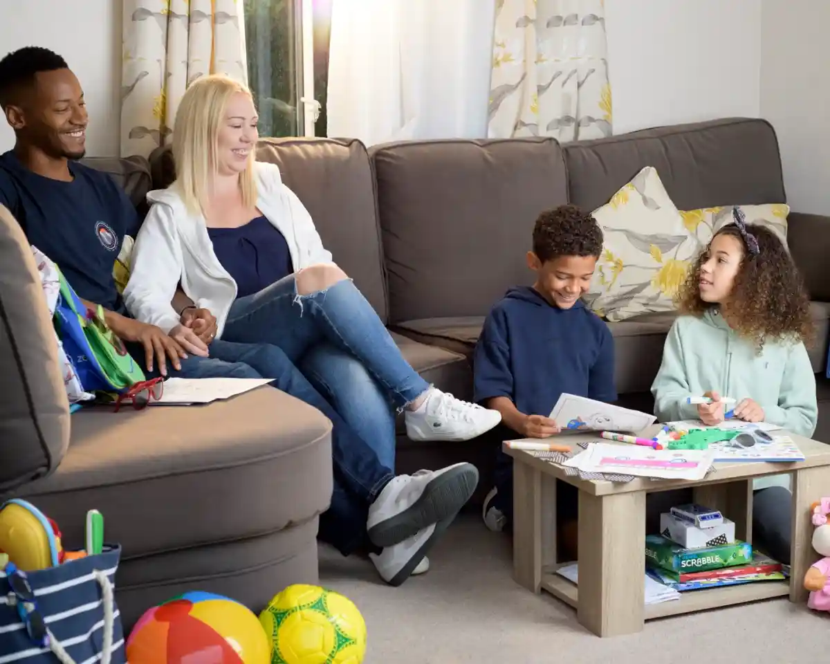 A family of four sits together in a cozy living room. An adult man and woman smile while seated on a couch, and two children are on the floor, engaged in a creative activity with coloring books and art supplies. Colorful toys are scattered around the room.