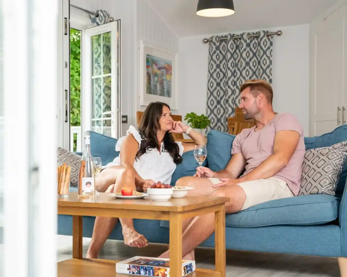 A couple sits on a blue couch in a bright, cozy living room, engaged in conversation. The woman is wearing a white top and has long dark hair, while the man is in a light pink shirt and shorts. A coffee table in front of them holds snacks and drinks. Sunlight streams in through nearby open doors.