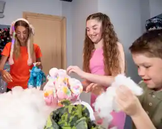 A girl with long, curly hair smiles while holding colorful soft toys, surrounded by fluffy cotton stuffing. Beside her, a boy plays with the cotton, and another girl in a bright outfit looks on, both engaged in a fun, creative activity. The setting appears to be a craft or playroom.