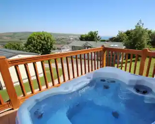 A hot tub with clear blue water is situated on a wooden deck, overlooking a scenic view of rolling hills and a distant coastline under a bright blue sky.