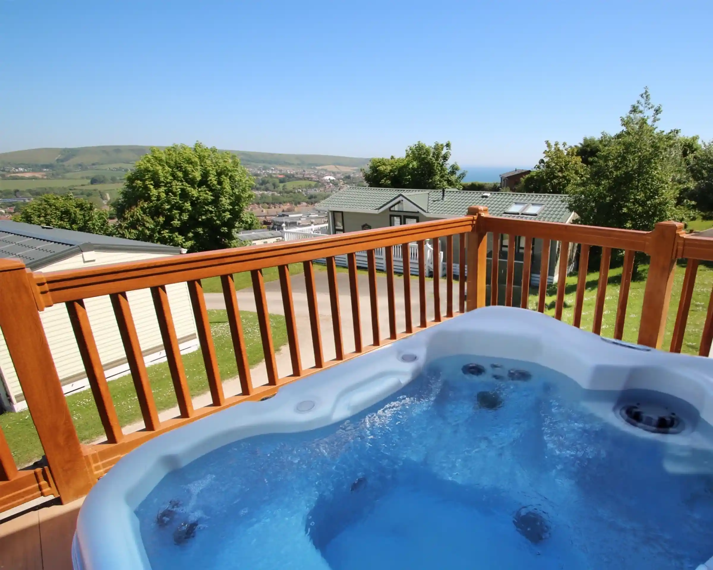 A hot tub with clear blue water is situated on a wooden deck, overlooking a scenic view of rolling hills and a distant coastline under a bright blue sky.