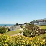 A scenic view of a coastal campsite featuring colourful flowers in the foreground, neatly arranged caravans on a hillside, and the ocean visible in the background under a clear blue sky.