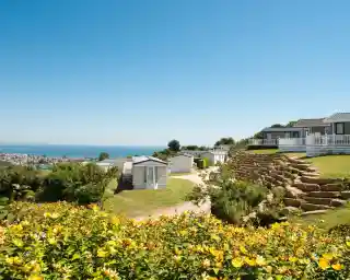 A scenic view of a coastal campsite featuring colourful flowers in the foreground, neatly arranged caravans on a hillside, and the ocean visible in the background under a clear blue sky.