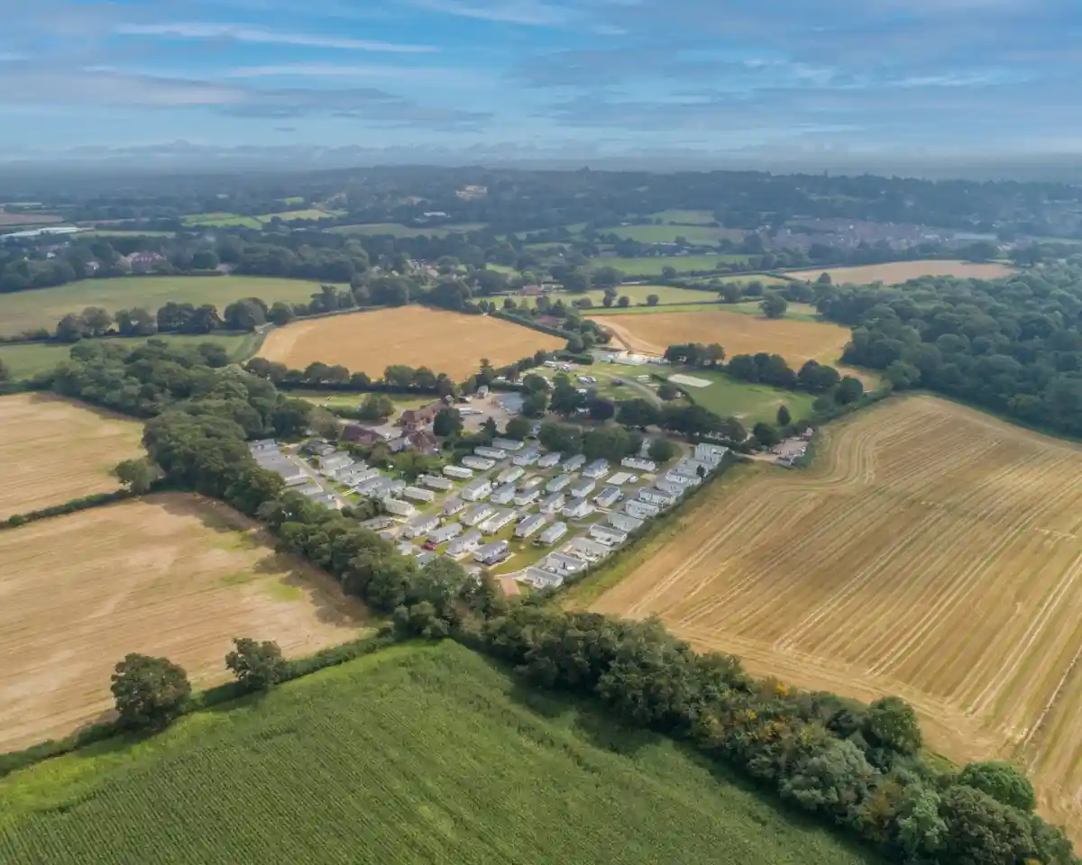 Aerial view of a rural landscape featuring farmland, green fields, and a cluster of mobile homes or caravans. The terrain includes patches of trees and a clear blue sky.