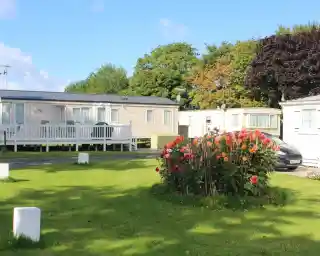 A green lawn with vibrant flower beds in the foreground, surrounded by several caravan-style homes under a clear blue sky. Trees in the background display autumn colors. A parked car is visible near the caravans.