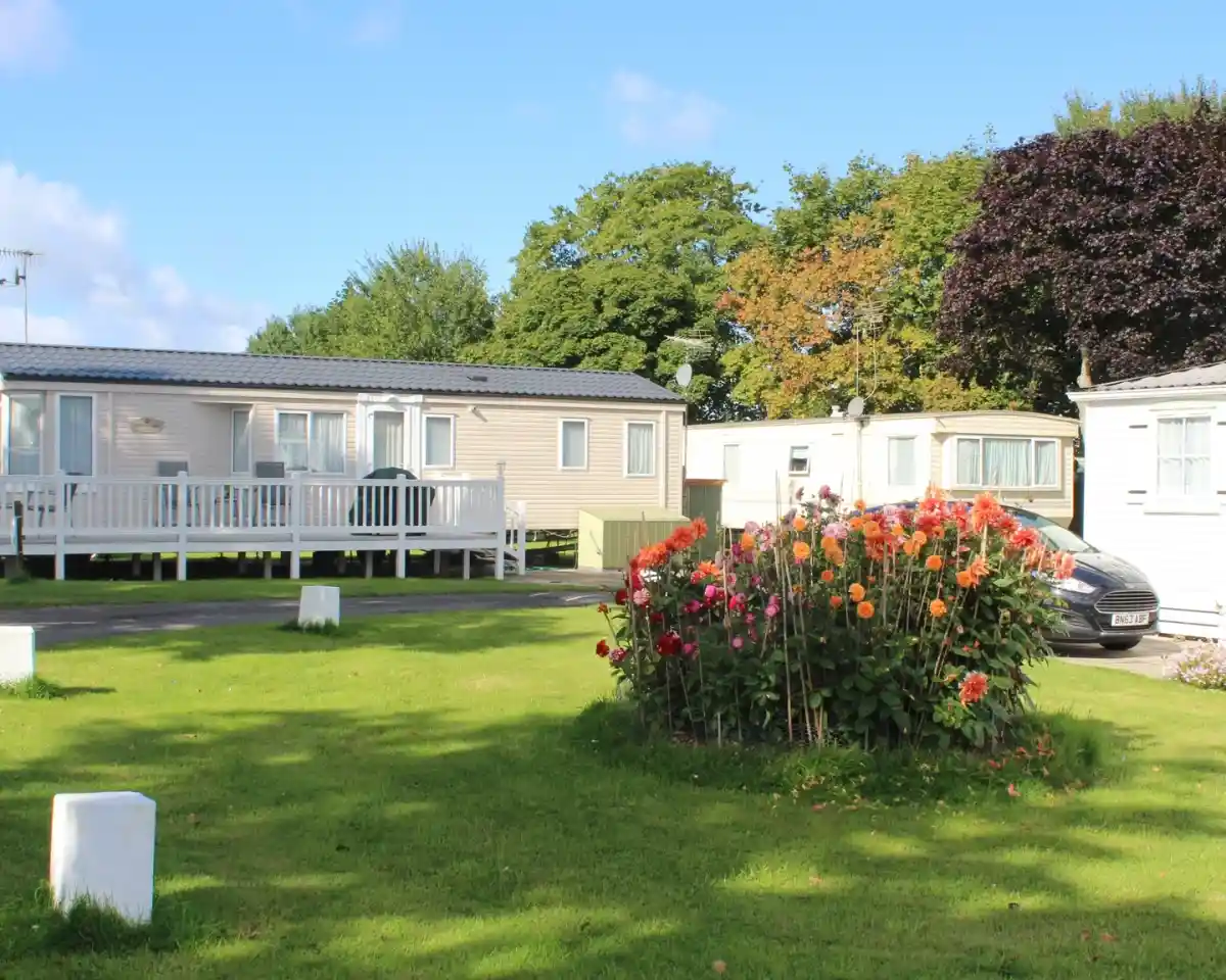 A green lawn with vibrant flower beds in the foreground, surrounded by several caravan-style homes under a clear blue sky. Trees in the background display autumn colors. A parked car is visible near the caravans.