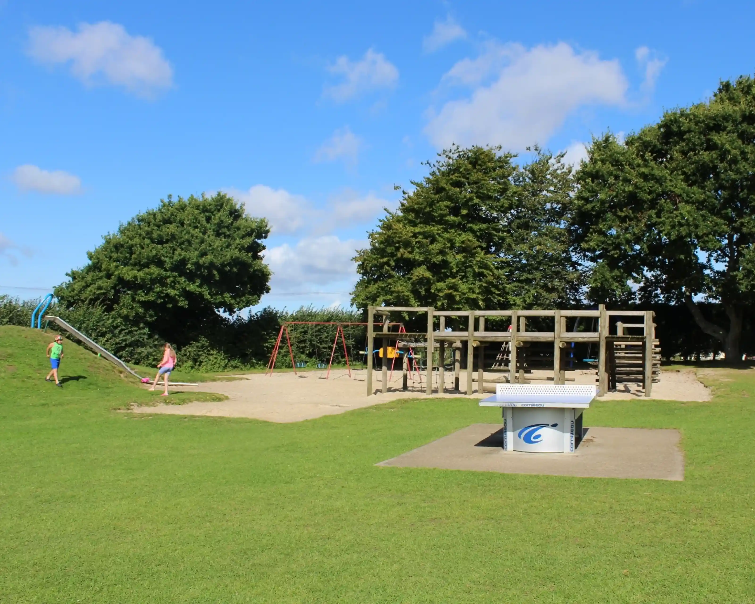 A children's playground featuring a wooden play structure, swings, and a slide, surrounded by grassy areas and trees under a blue sky with fluffy clouds.