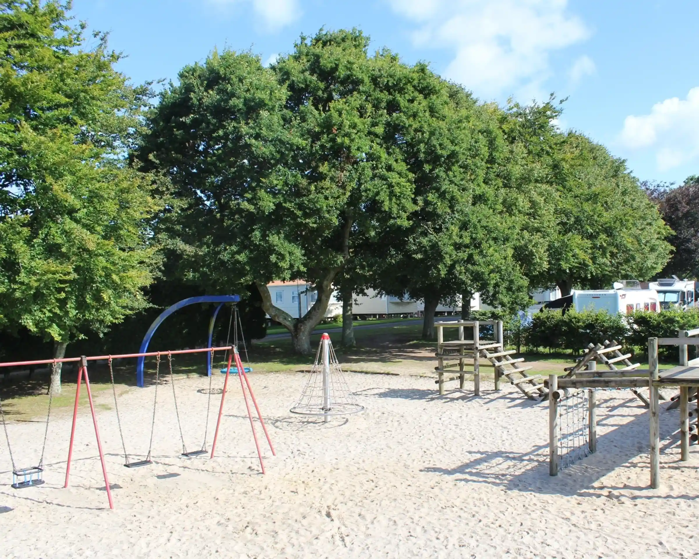 A playground featuring swings, a climbing structure, and a large sandbox, surrounded by trees and residential structures in the background. The sky is clear with a few clouds.