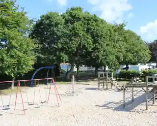 A playground featuring swings, a climbing structure, and a large sandbox, surrounded by trees and residential structures in the background. The sky is clear with a few clouds.