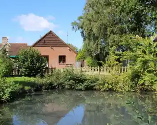 A quaint brick house with a sloped roof sits beside a tranquil pond surrounded by lush greenery. Clear blue skies and fluffy clouds can be seen overhead.