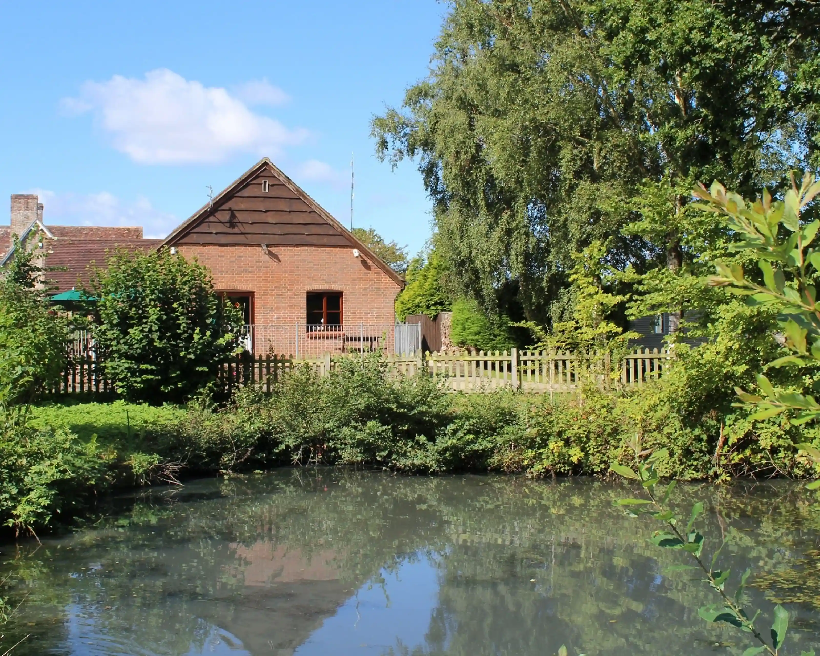 A quaint brick house with a sloped roof sits beside a tranquil pond surrounded by lush greenery. Clear blue skies and fluffy clouds can be seen overhead.